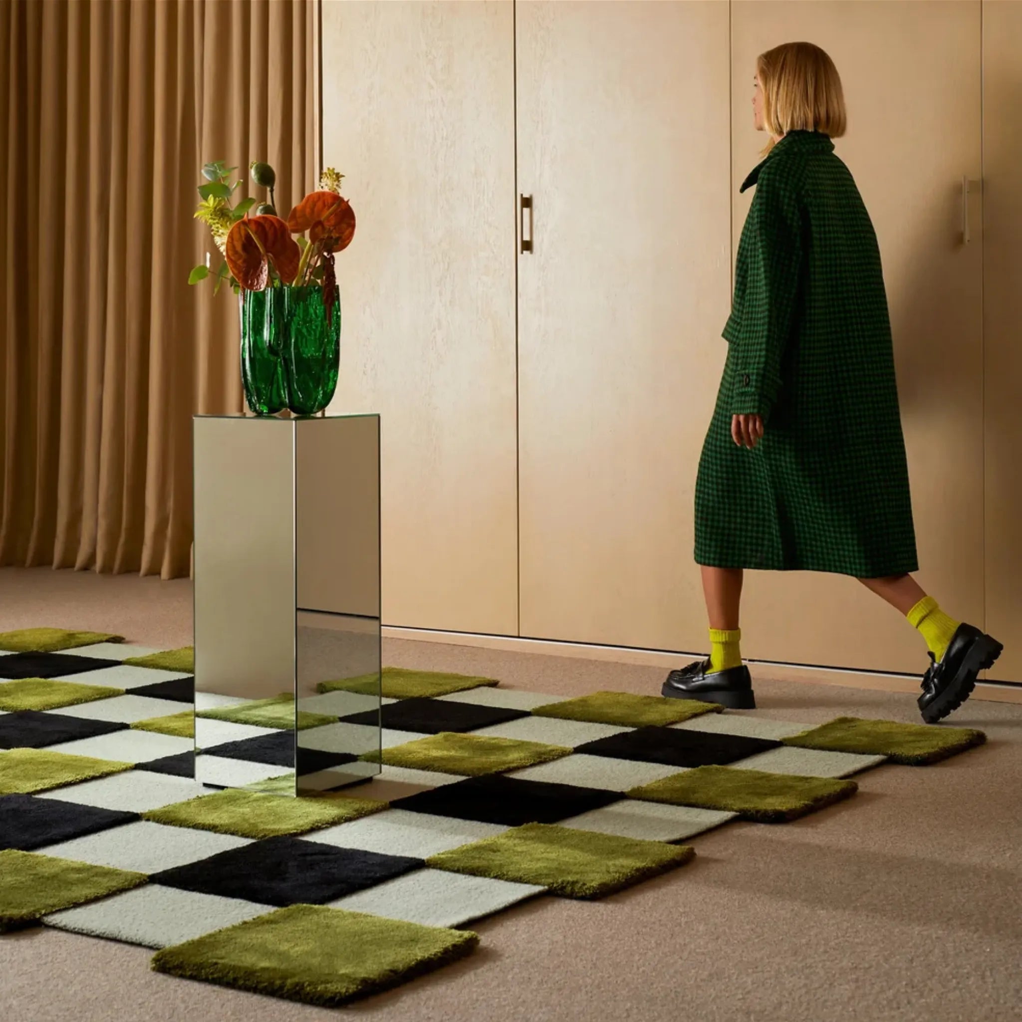 Woman in a green coat standing on a Green Black Mint Checkered Irregular High Low Rug with a mirror and flowers.