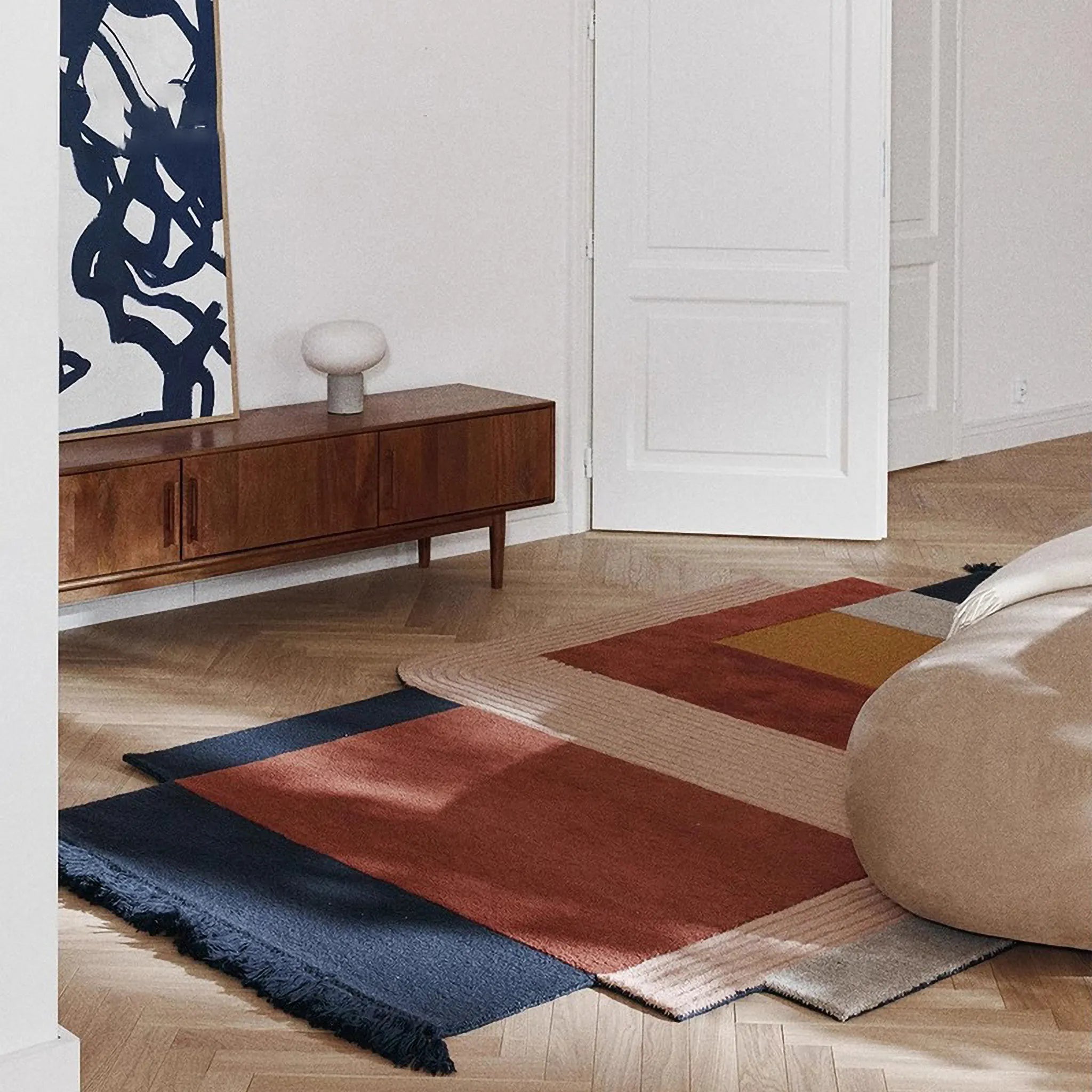 Rust Mustard Navy Pink Abstract Irregular Fringe Wool Rug on a wooden floor with a wooden cabinet and a wall art in the background.