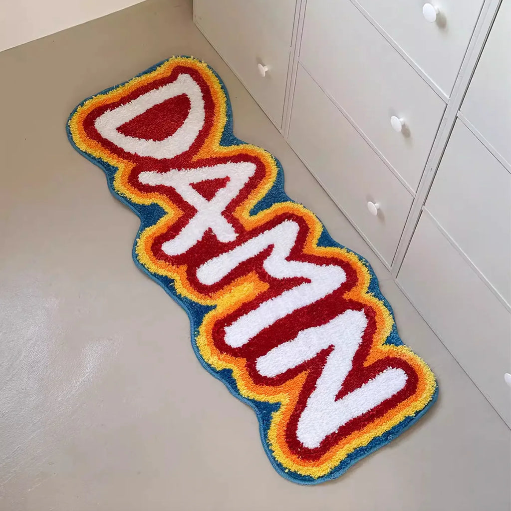 A hand-tufted rainbow coloured runner rug by Housenfriends with the word 'DAMN' on a light-colored floor.