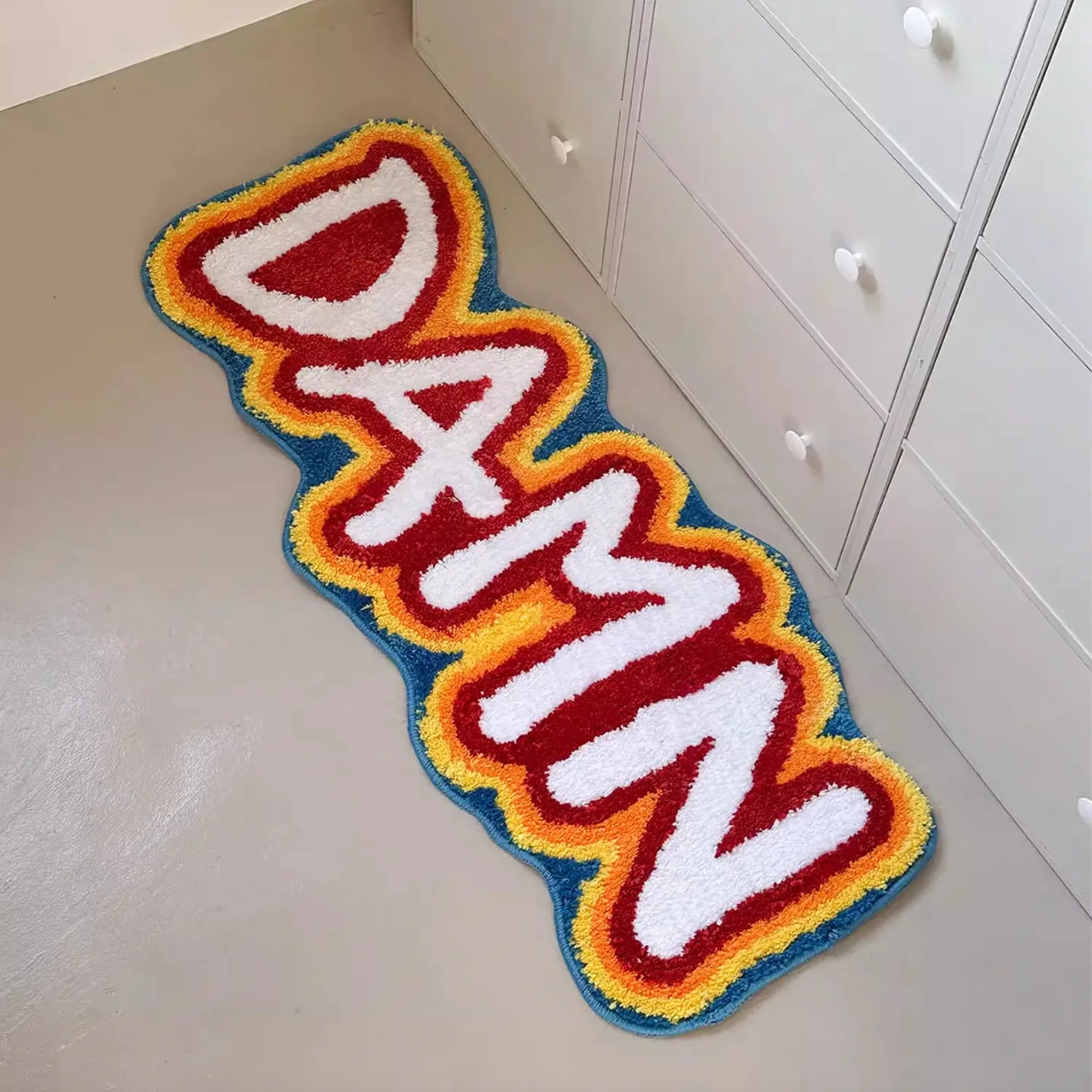 A hand-tufted rainbow coloured runner rug by Housenfriends with the word 'DAMN' on a light-colored floor.