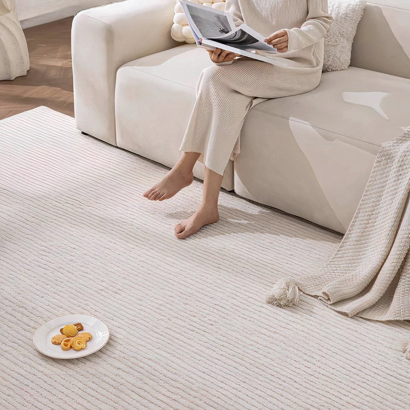 Person on a beige sofa reading on Rae off-white minimalist rug by Housenfriends with cookies nearby.