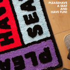 Corner of 'Please Have a Seat' graphic word art rug in purple, black, pink and blue by Housenfriends on a wooden floor.