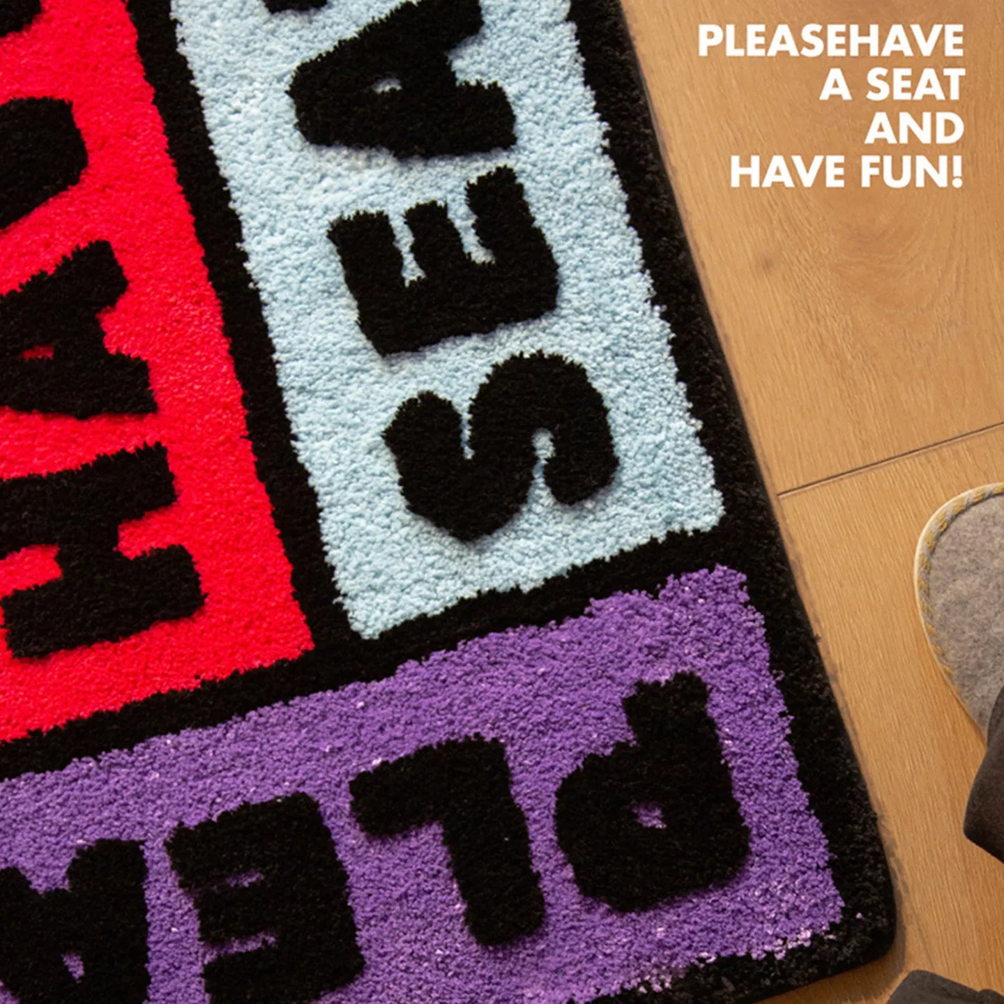 Corner of 'Please Have a Seat' graphic word art rug in purple, black, pink and blue by Housenfriends on a wooden floor.