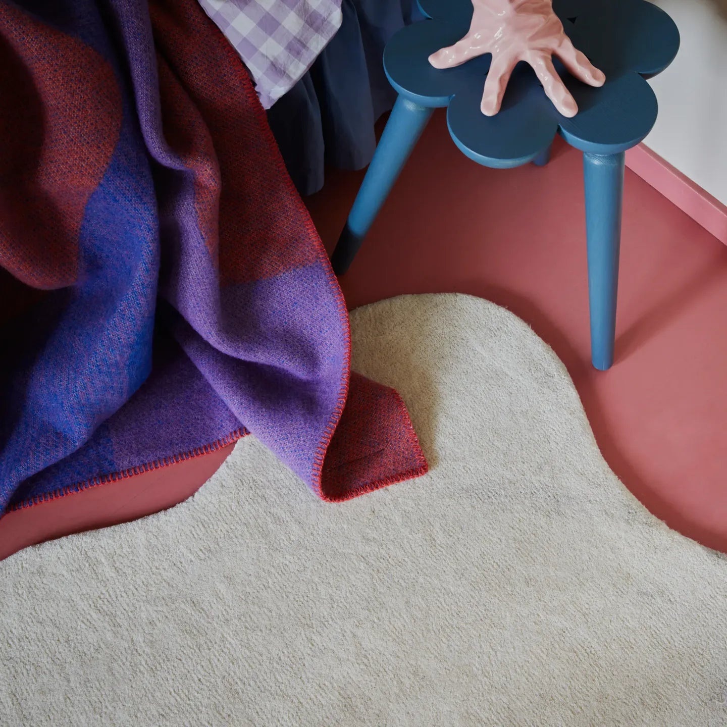 A corner of the Cora off-white irregular wavy rug by Housenfriends with a blanket on top, on red floor with blue stool