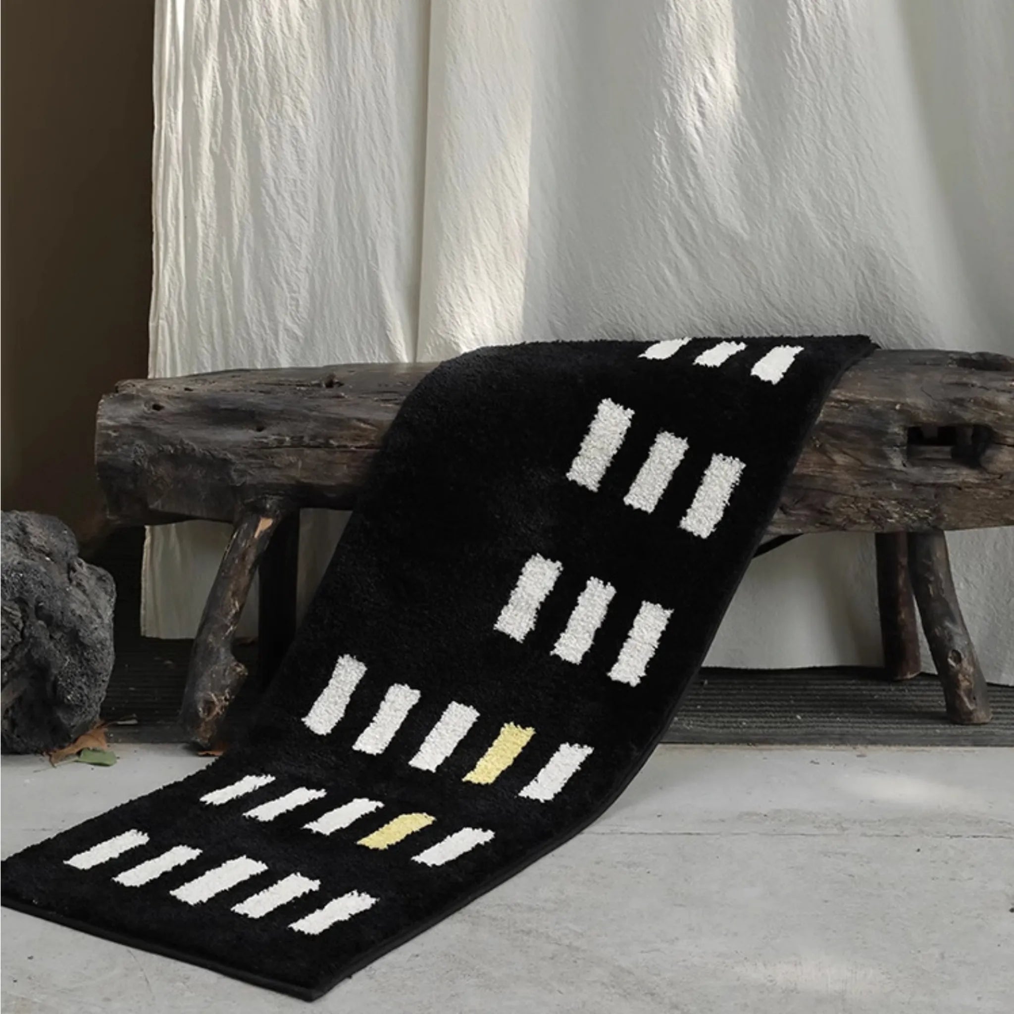 Black rug with white and yellow pattern draped over a wooden bench against a white curtain background.