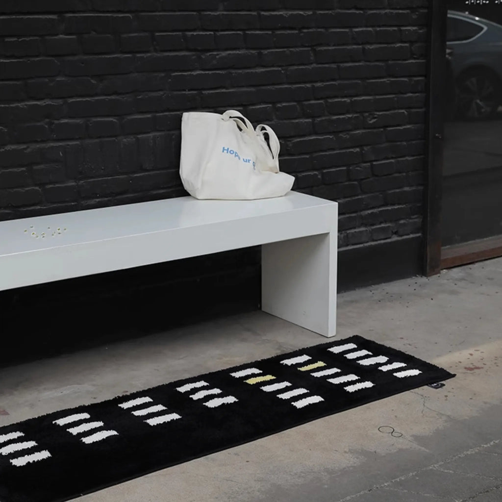 White bench with a white bag against a black brick wall, Arlo Black and White Asymmetric Checkered Tufted Runner Rug on the floor.