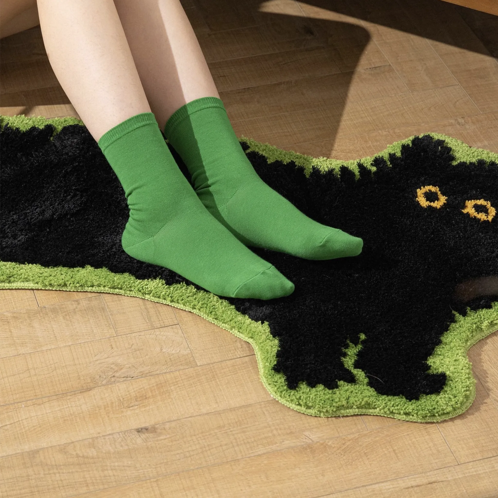 Person wearing green socks stepping onto a black arched cat-shaped rug with green border on a wooden floor.