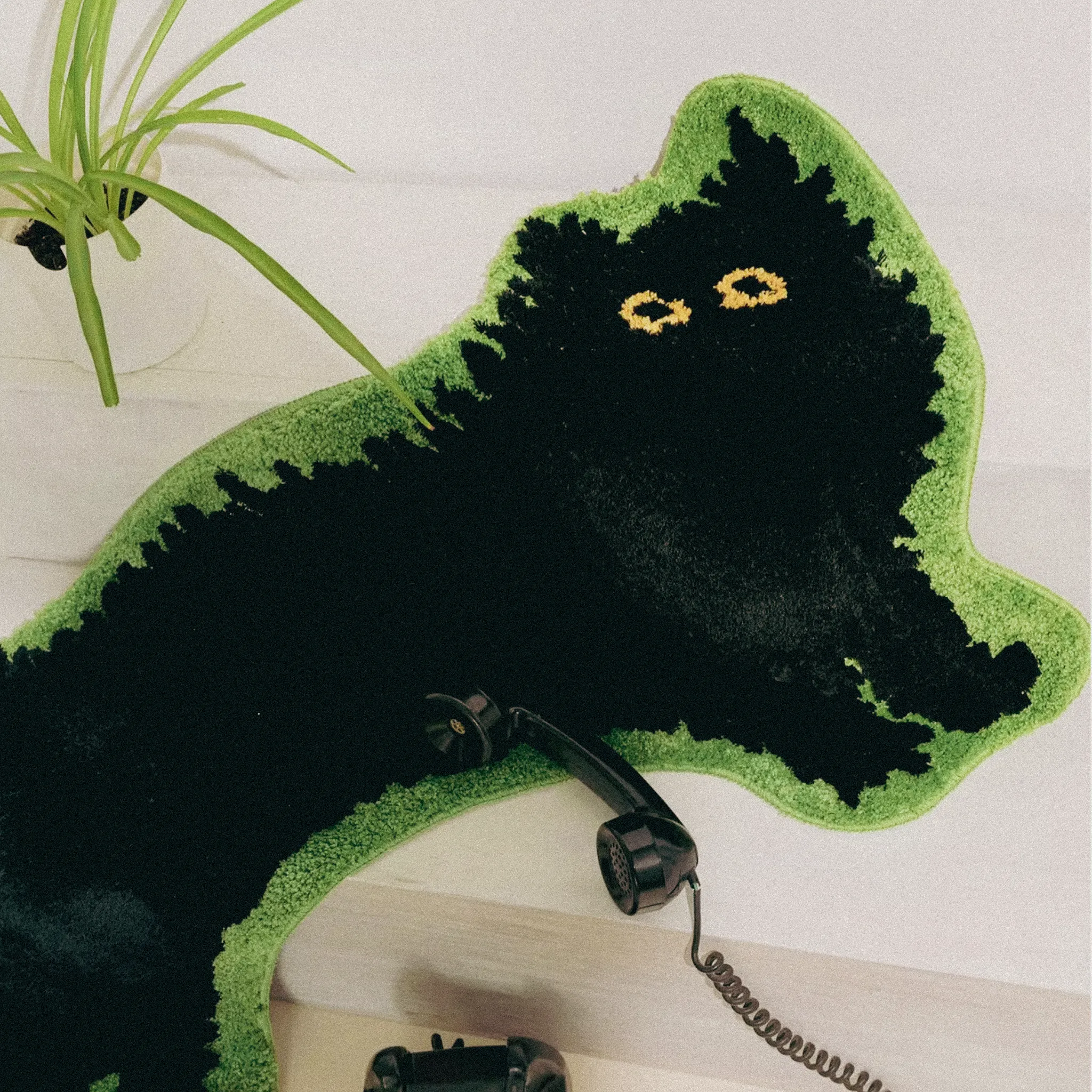 Black arched cat-shaped tufted rug with green border on a light wooden floor with a plant.