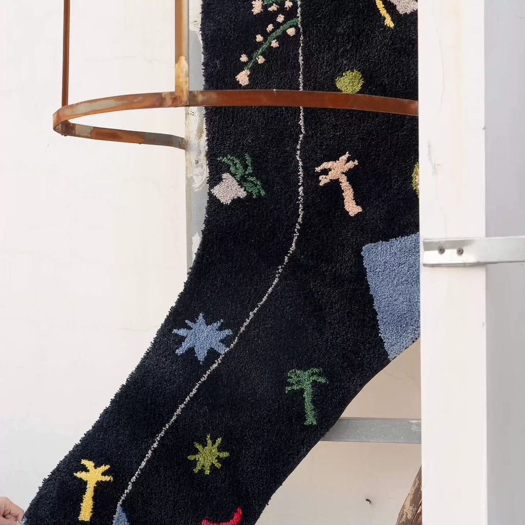 Black graphic motif sock shaped rug with raised flower, palm and starburst details hanging over a rail by Housenfriends