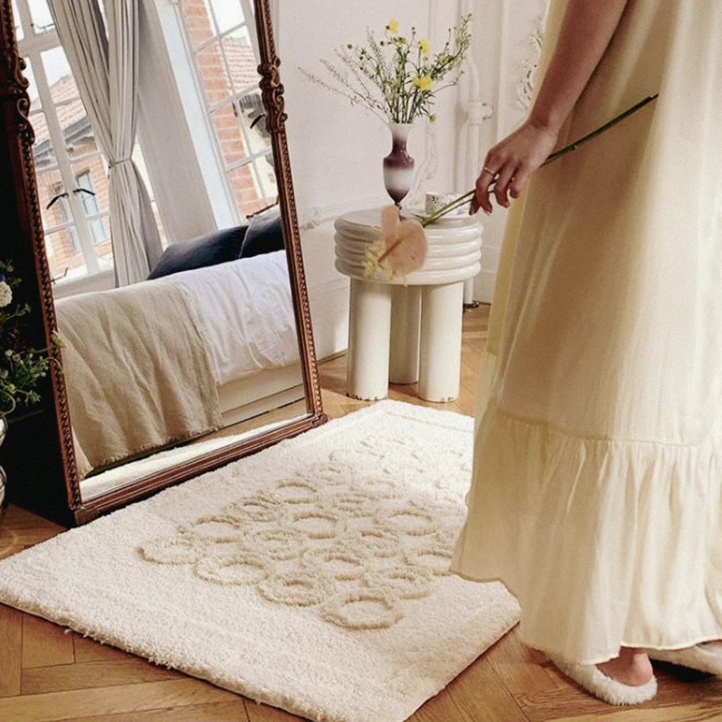 Woman in a yellow dress standing next to a large mirror on Nola Cream Beige Sculpted Circle Tufted Rug.