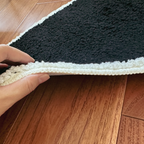 Close-up of a hand holding the corner of a black and white textured rug on a wooden floor to demonstrate its thick pile.