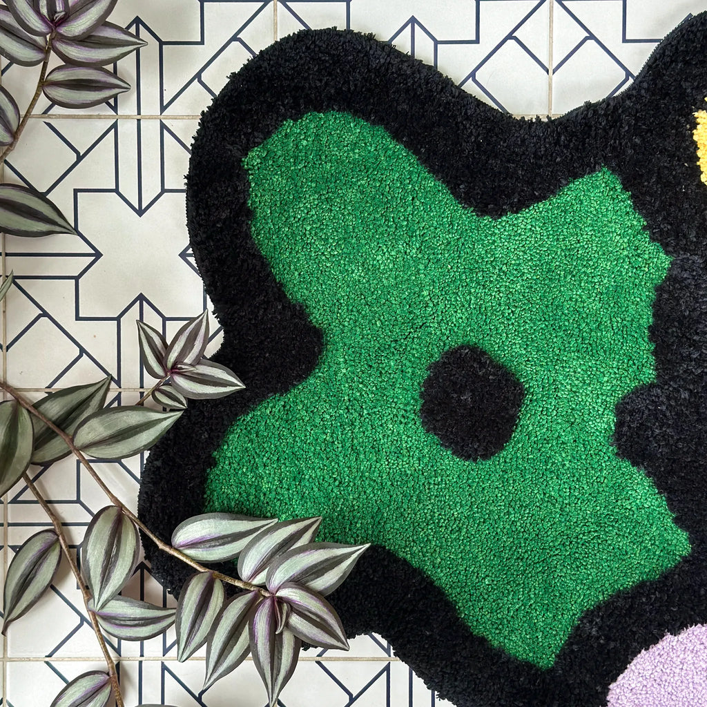 A close-up of a plush, high-pile shag rug with an organic, abstract shape. The rug features a vibrant green center, a thick black border, and a small black circle in the middle.
