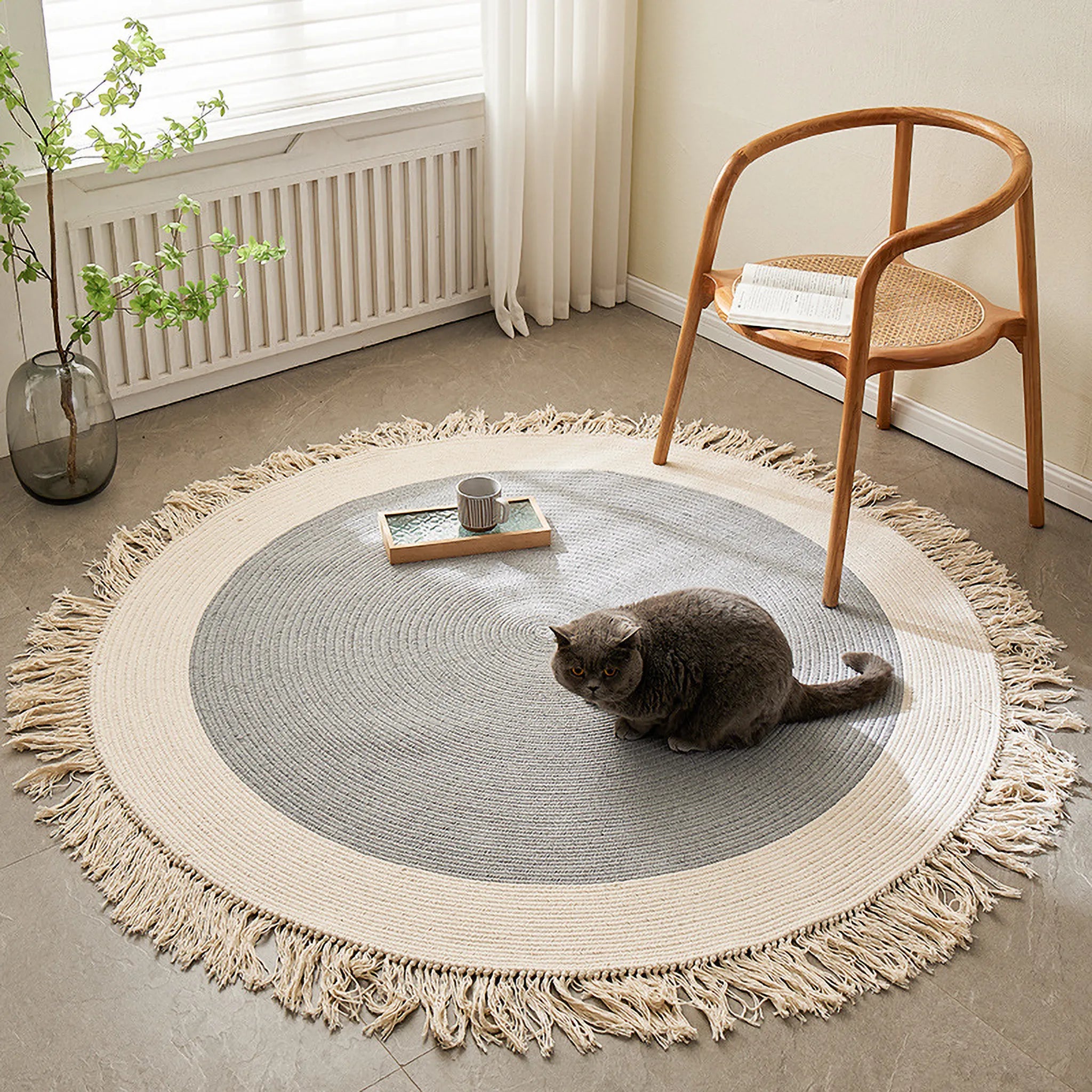 Signe grey and natural fringed round hand-braided cotton blend rug styled in a bright room corner with chair and cat by Housenfriends