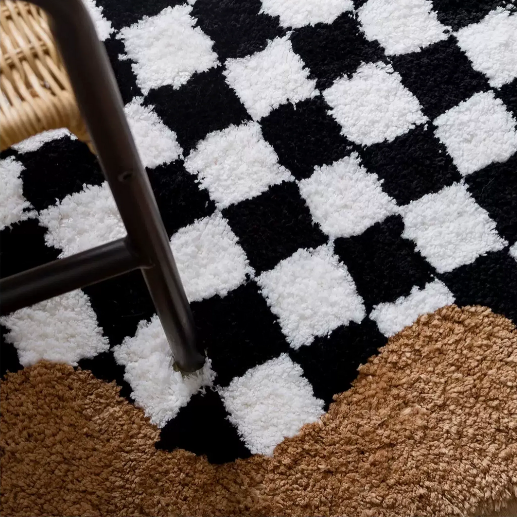 A close-up of Tess Black White Retro Checkered Bronze Wavy Edge Tufted Rug, with part of a dark chair leg visible.