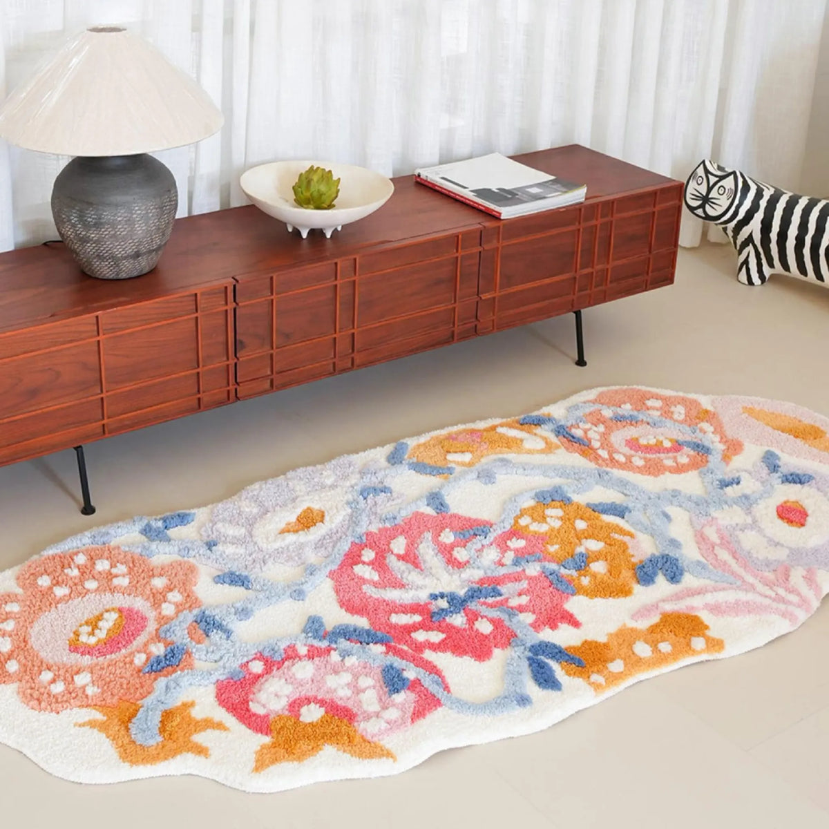 Celia irregular floral runner rug in cream, coral pink, orange and powder blue, styled beside a long wooden sideboard with a table lamp and striped cat ornament.