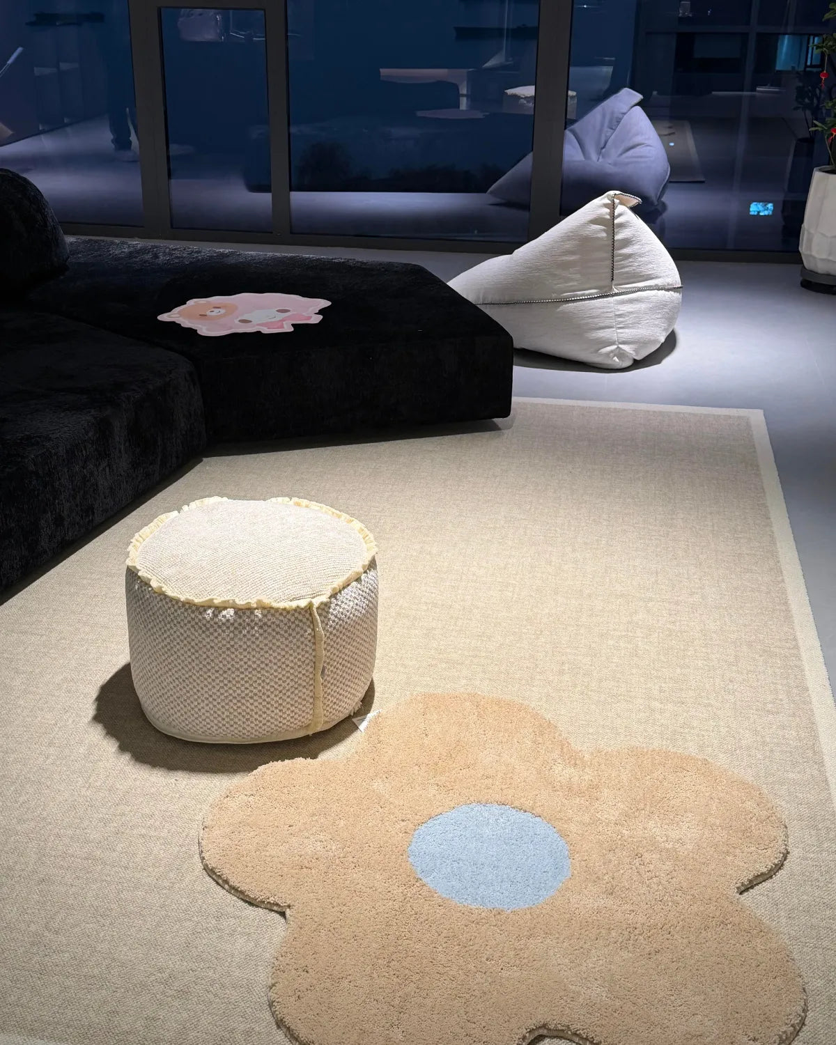 Housenfriends Maisie cream and pale blue flower-shaped rug, 100 × 100 cm, layered over a neutral rug in front of a low sofa and beside a pouf.
