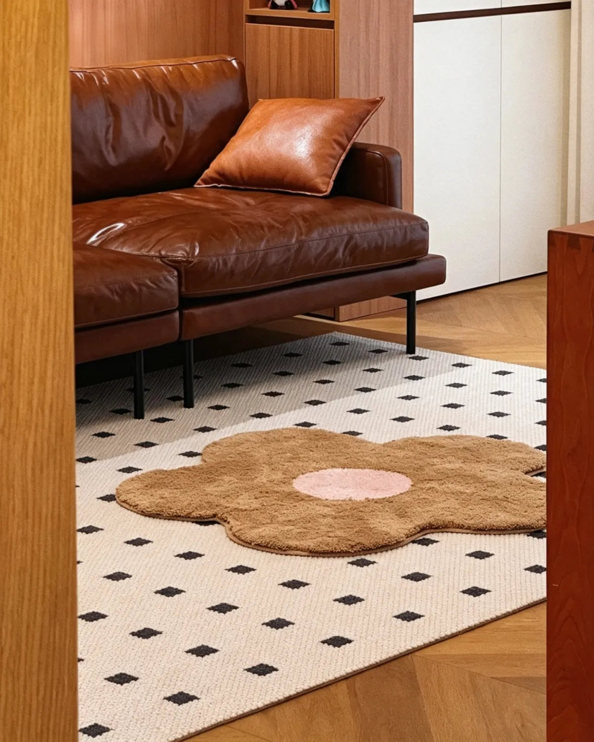 Housenfriends Maisie beige and blush flower-shaped rug, 100 × 100 cm, placed on a cream patterned rug in front of a brown leather sofa.