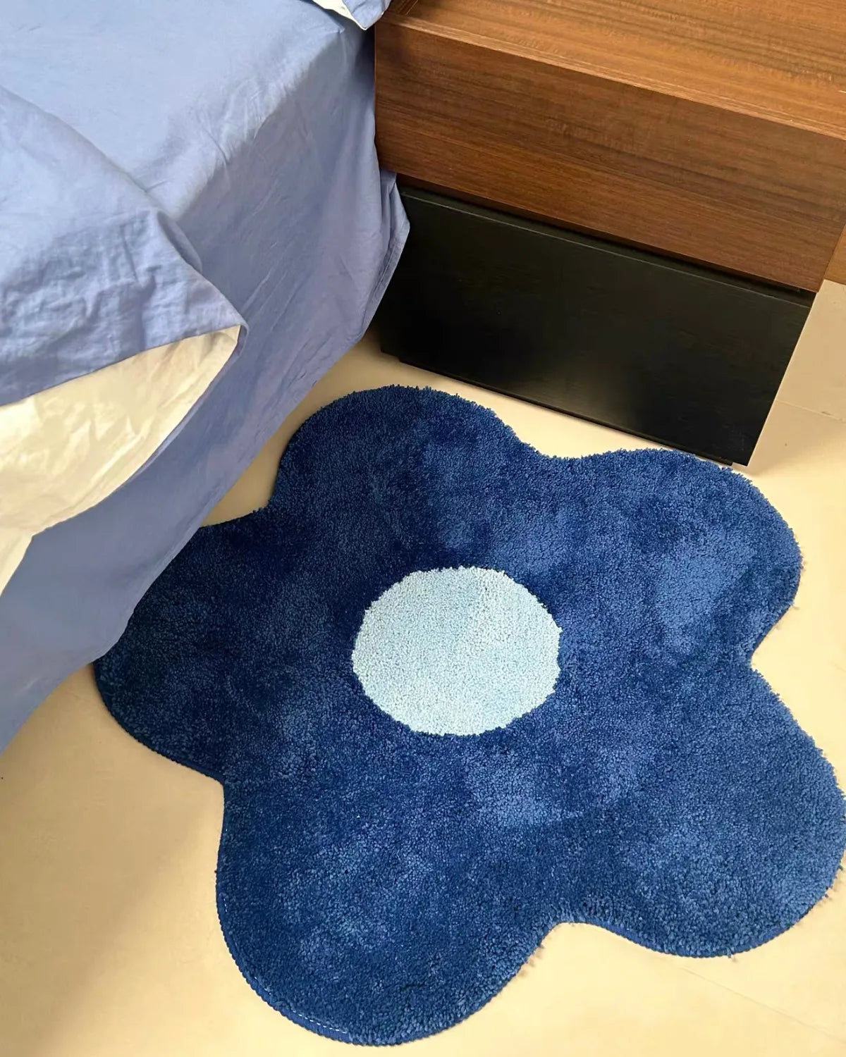 Housenfriends Maisie blue and pale blue daisy-shaped rug, 80 × 80 cm, beside a blue bed and wooden bedside table.