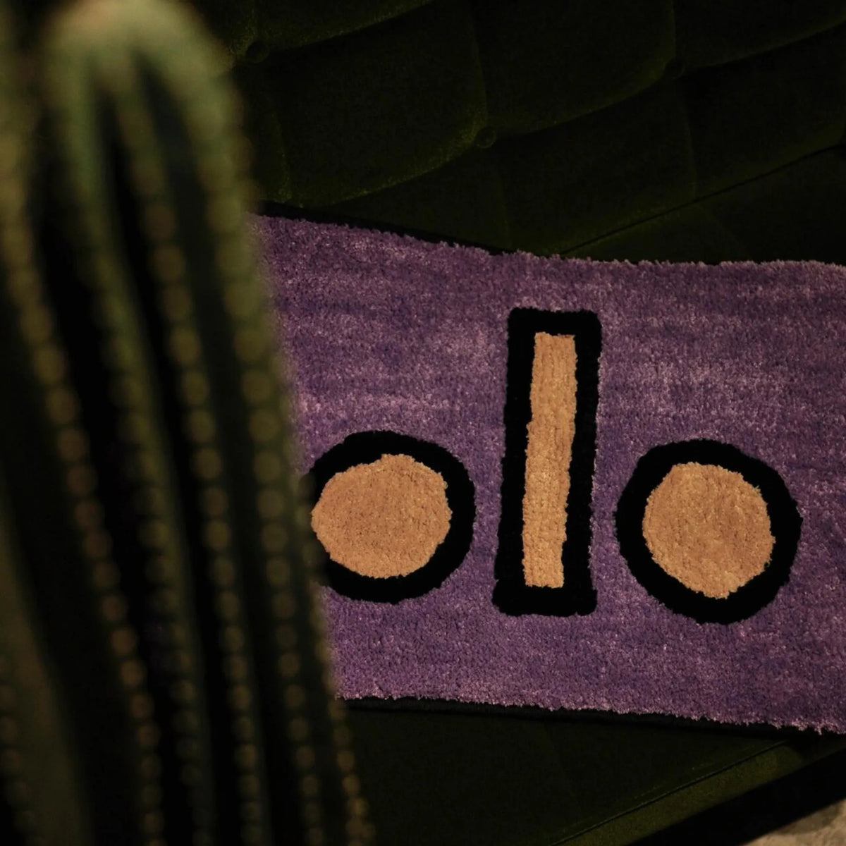 Housenfriends Nico purple abstract male body art rug draped over a dark green velvet sofa, with a cactus in the foreground.