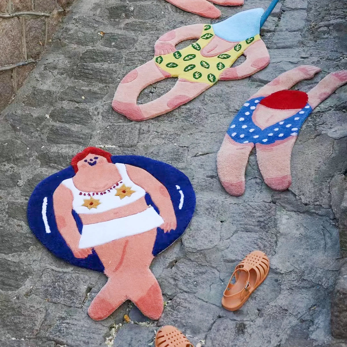 Three playful swimming girl body art rugs displayed on stone paving, featuring Housenfriends' June in a white swimsuit and navy blue swim ring.