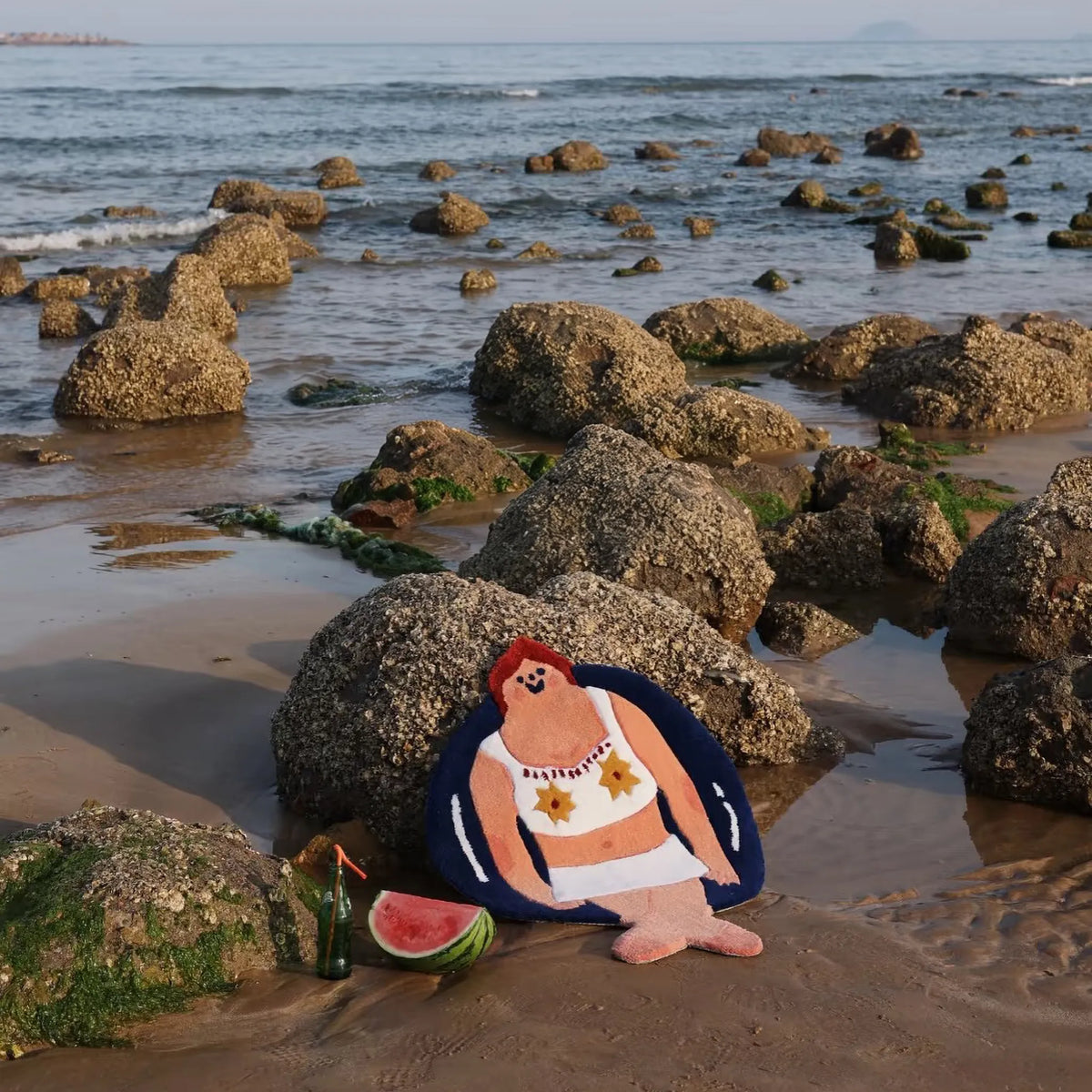Housenfriends June swimming girl body art rug resting against coastal rocks on a sandy beach, with the sea in the background.